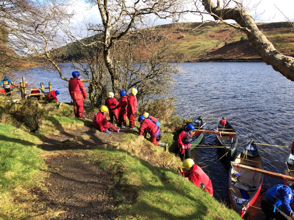 A large group of people in red safety suits and blue and yellow helmets climbing out of canoes onto a river bank