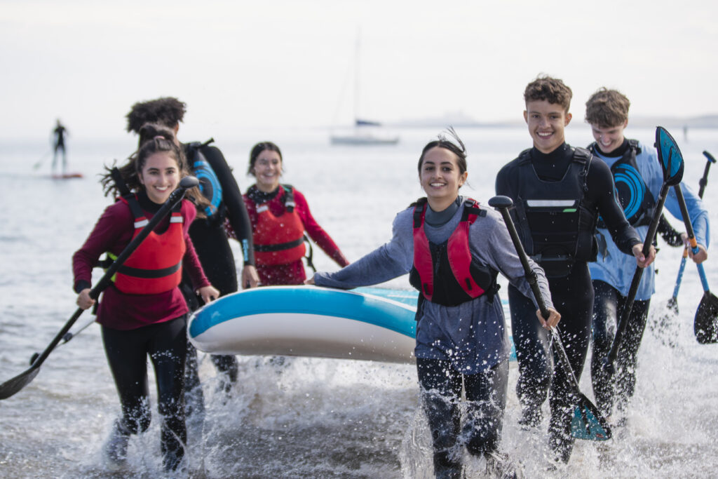 Group of mixed ethnic and age teens running in the ocean while carrying a multiple person paddle board together in the North East of England.