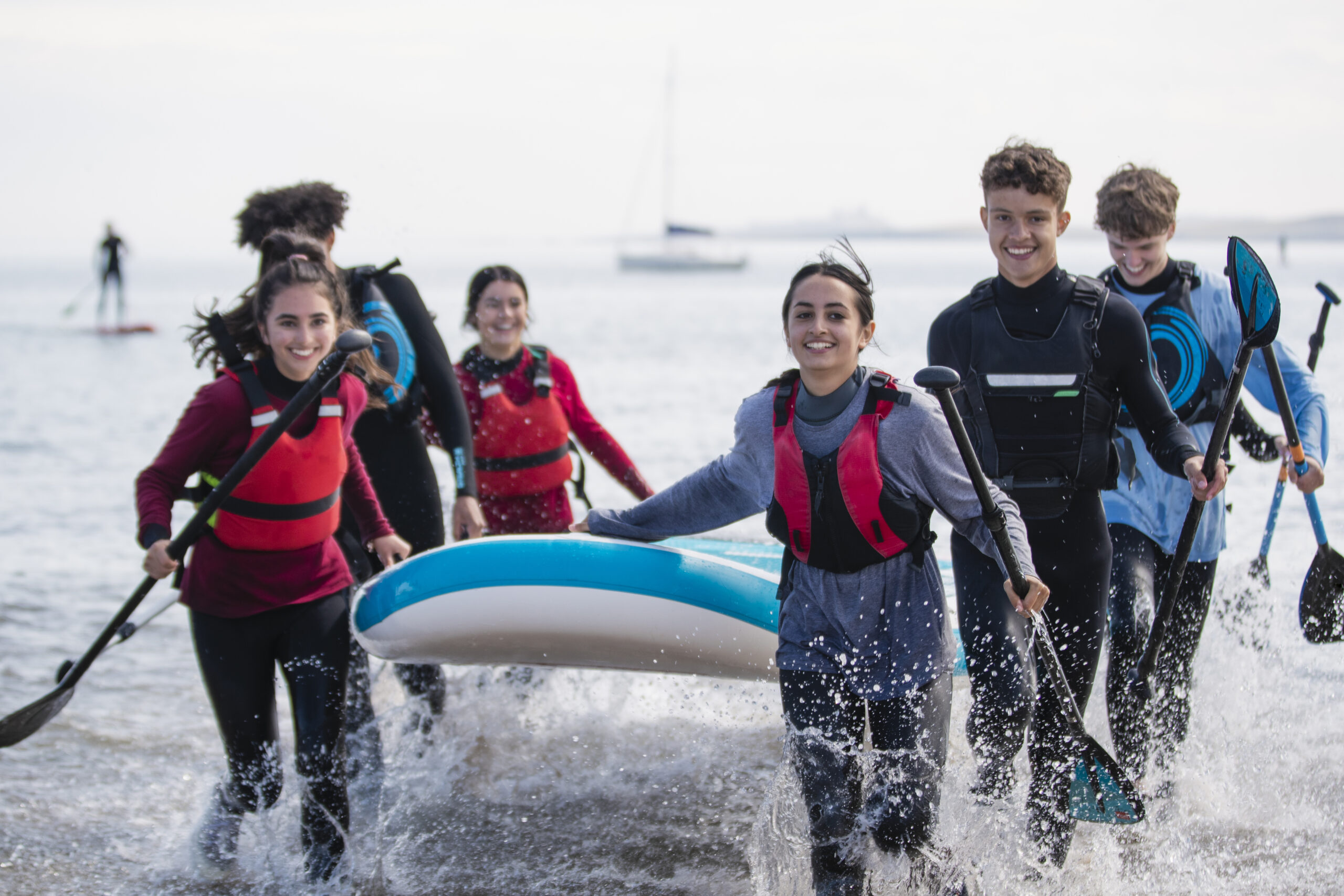 Group of mixed ethnic and age teens running in the ocean while carrying a multiple person paddle board together in the North East of England.