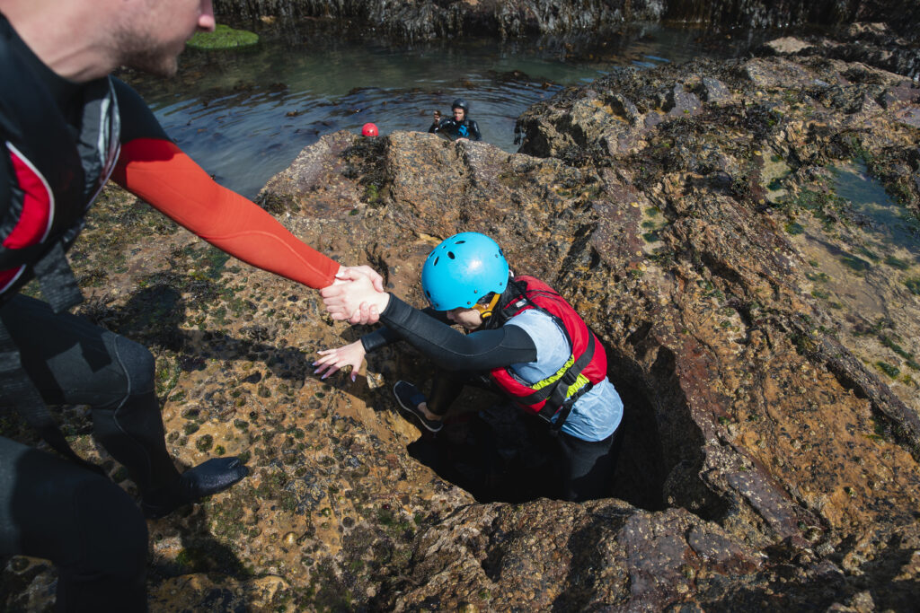 Group of mixed ethnic friends coasteering in the North East of England at Beadnell. They are wearing helmets and life jackets and one man is helping a young person climbing through a hole of a cave while they explore.